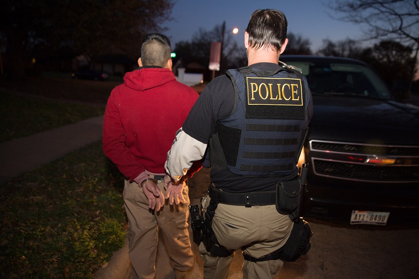 Officer in a police vest escorts a handcuffed man beside a vehicle