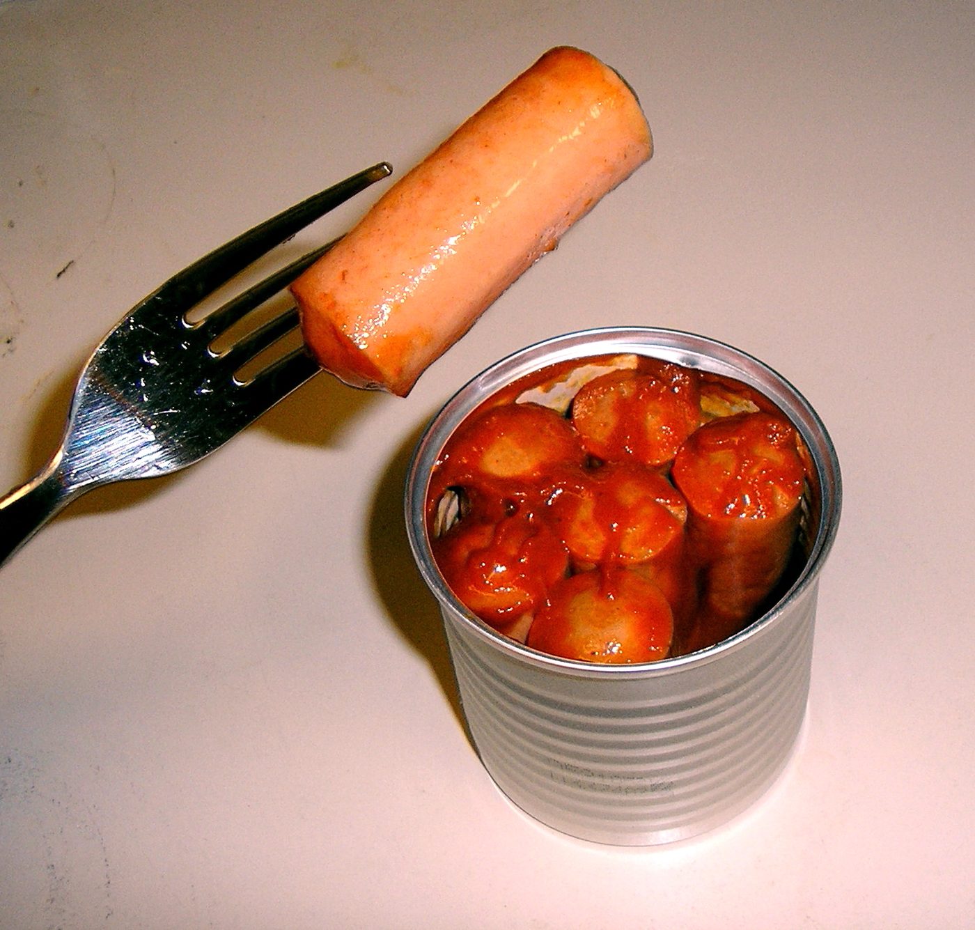 An open can of Vienna sausages in barbecue sauce, with one sausage on a fork