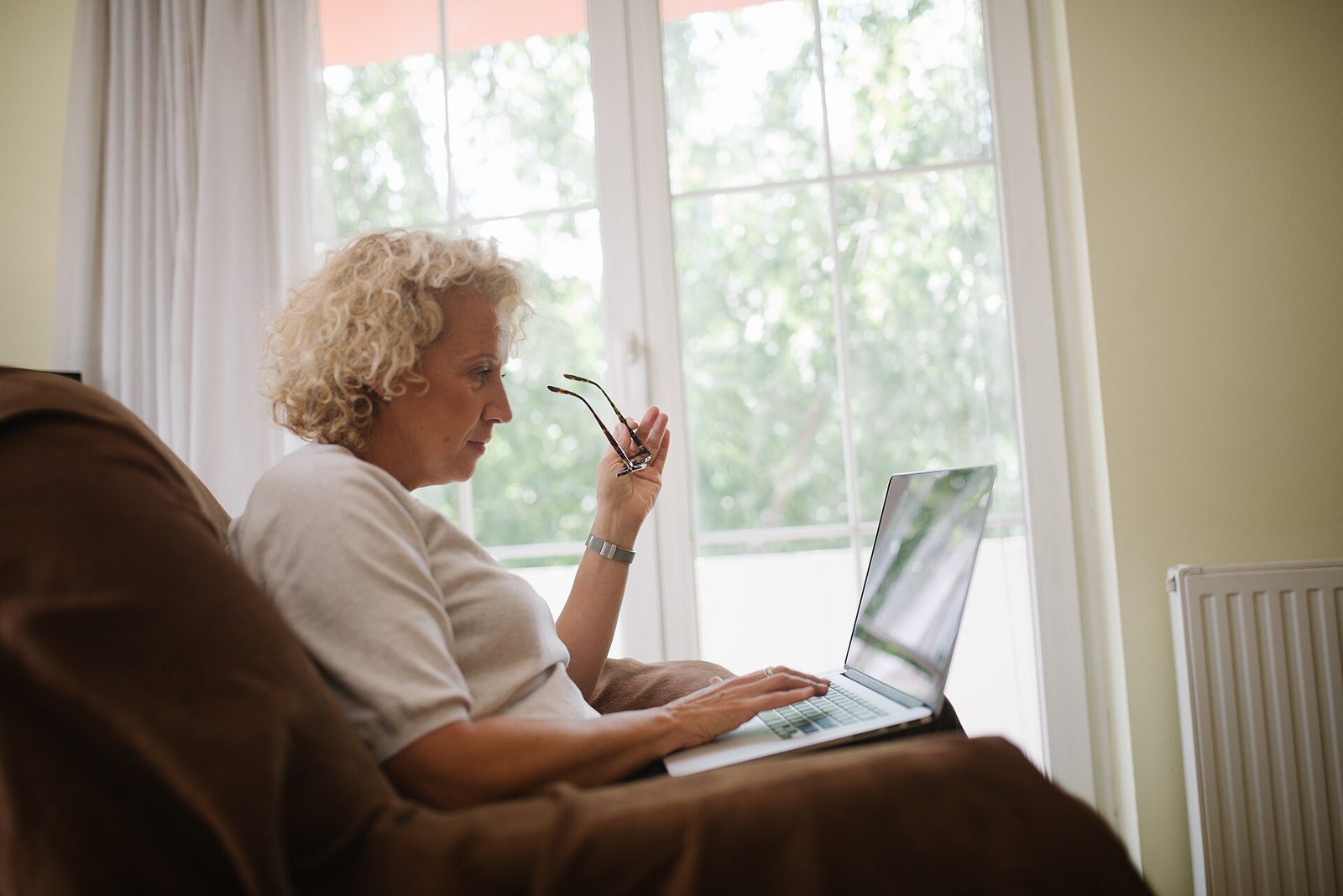 An older woman holds her glasses while typing on a laptop in an armchair