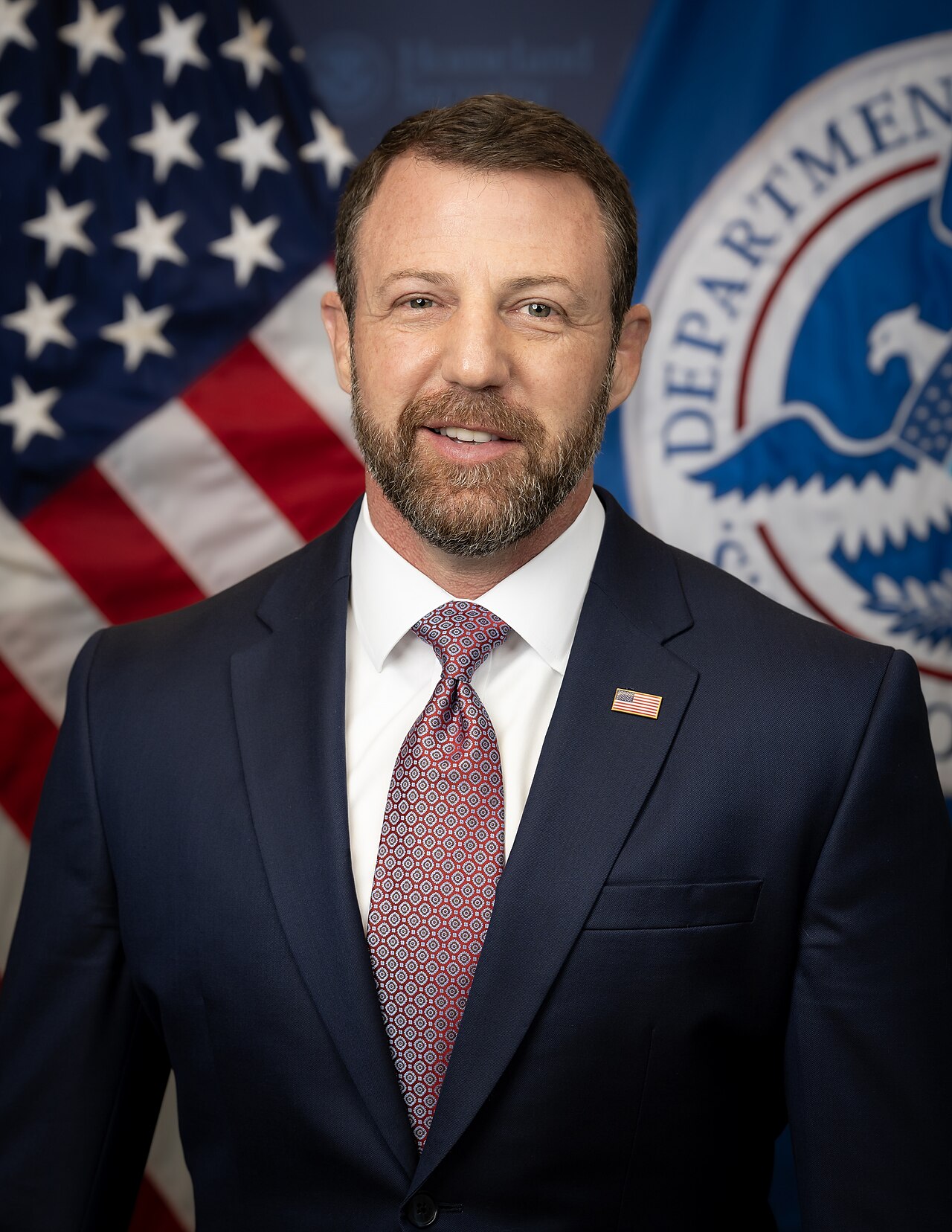 Official portrait of Homeland Security Secretary Markwayne Mullin in front of U.S. and DHS flags, March 2026