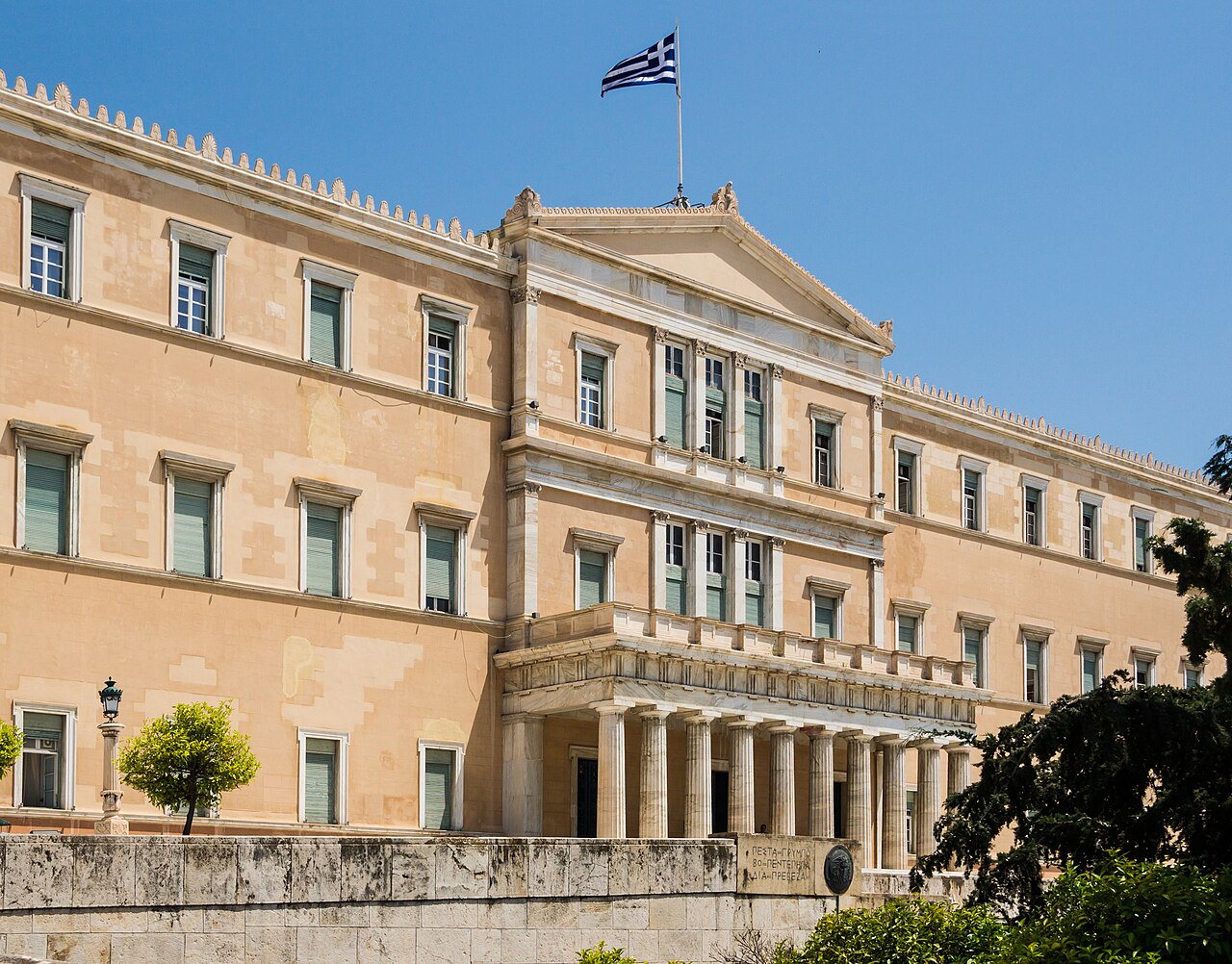 The front of the Hellenic Parliament building in Athens under a clear blue sky, with a Greek flag flying above it