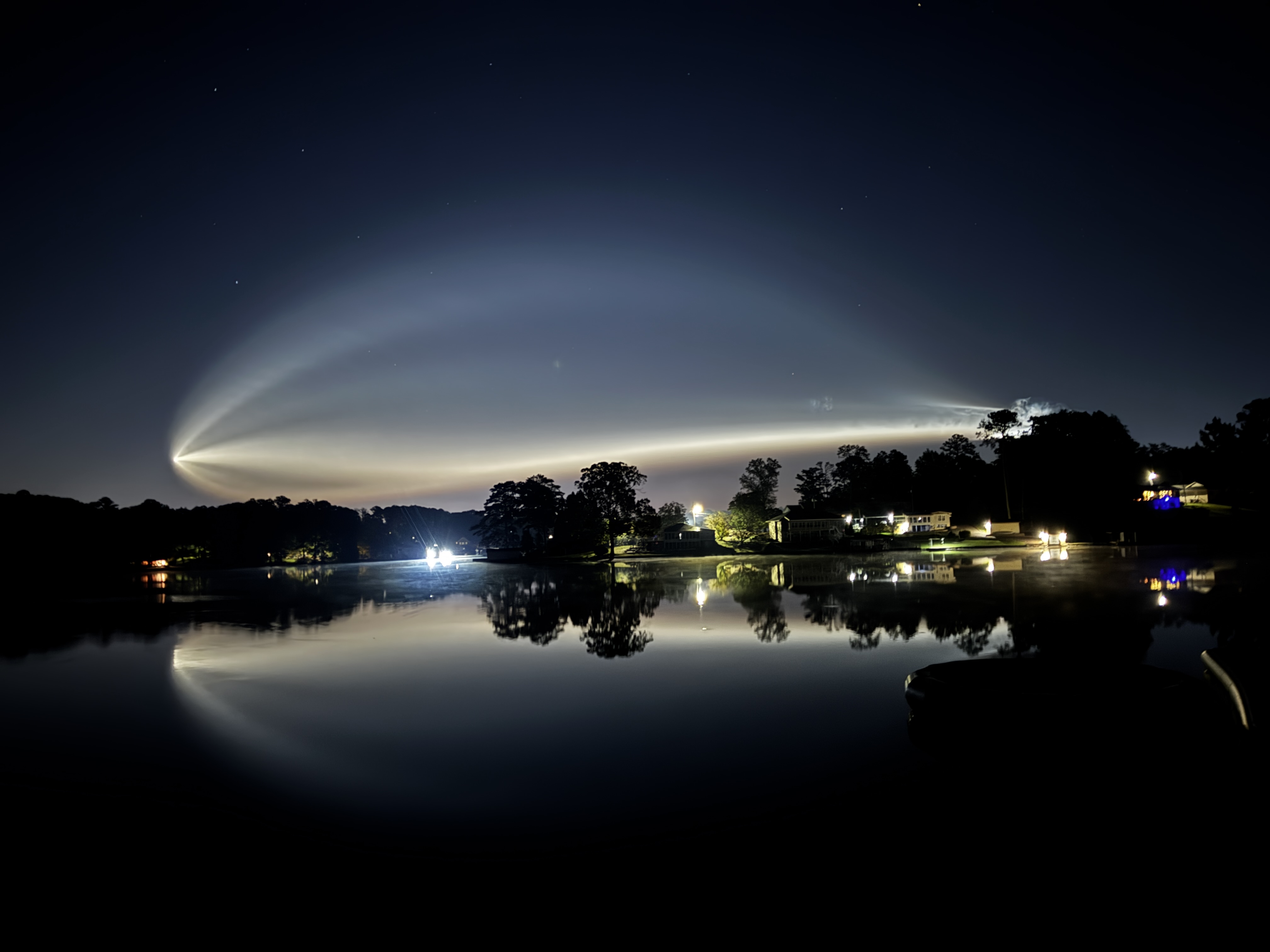 The exhaust plume from a Falcon 9 launch spreads across the predawn sky, reflected in a calm lake.