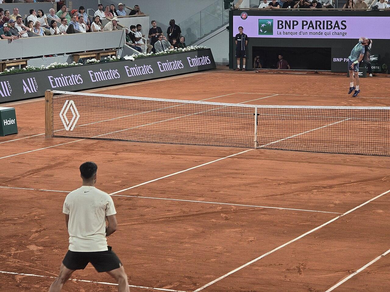 Carlos Alcaraz, in the foreground, waits to return a serve from Damir Džumhur on a clay court at the 2025 French Open