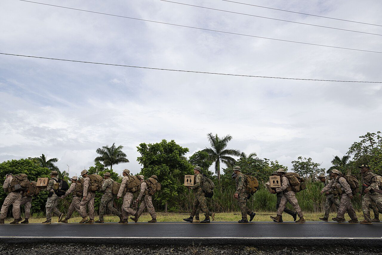 A line of soldiers in camouflage with backpacks walking along a road past palm trees