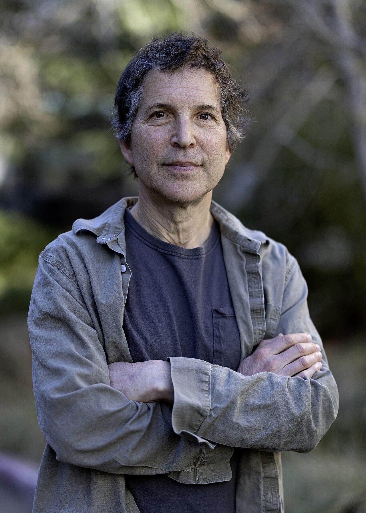 David Baker, a man with curly dark hair, stands with his arms crossed in front of green trees