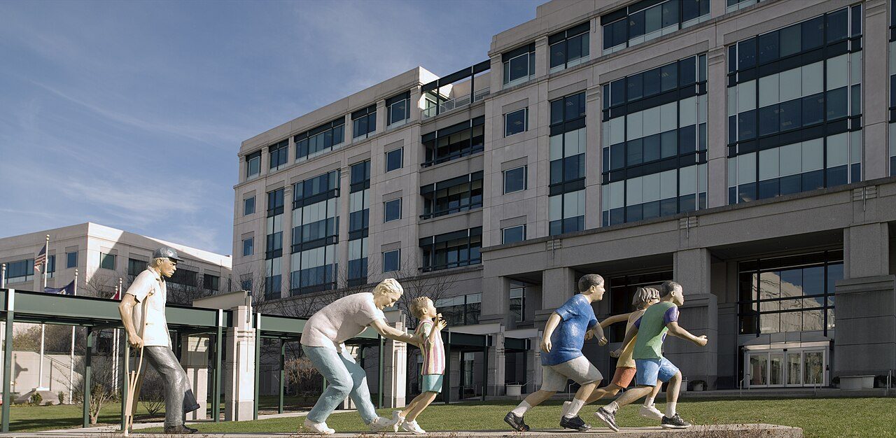 Painted sculptures of a man on crutches, a woman with a child and running children on a lawn outside a large office building