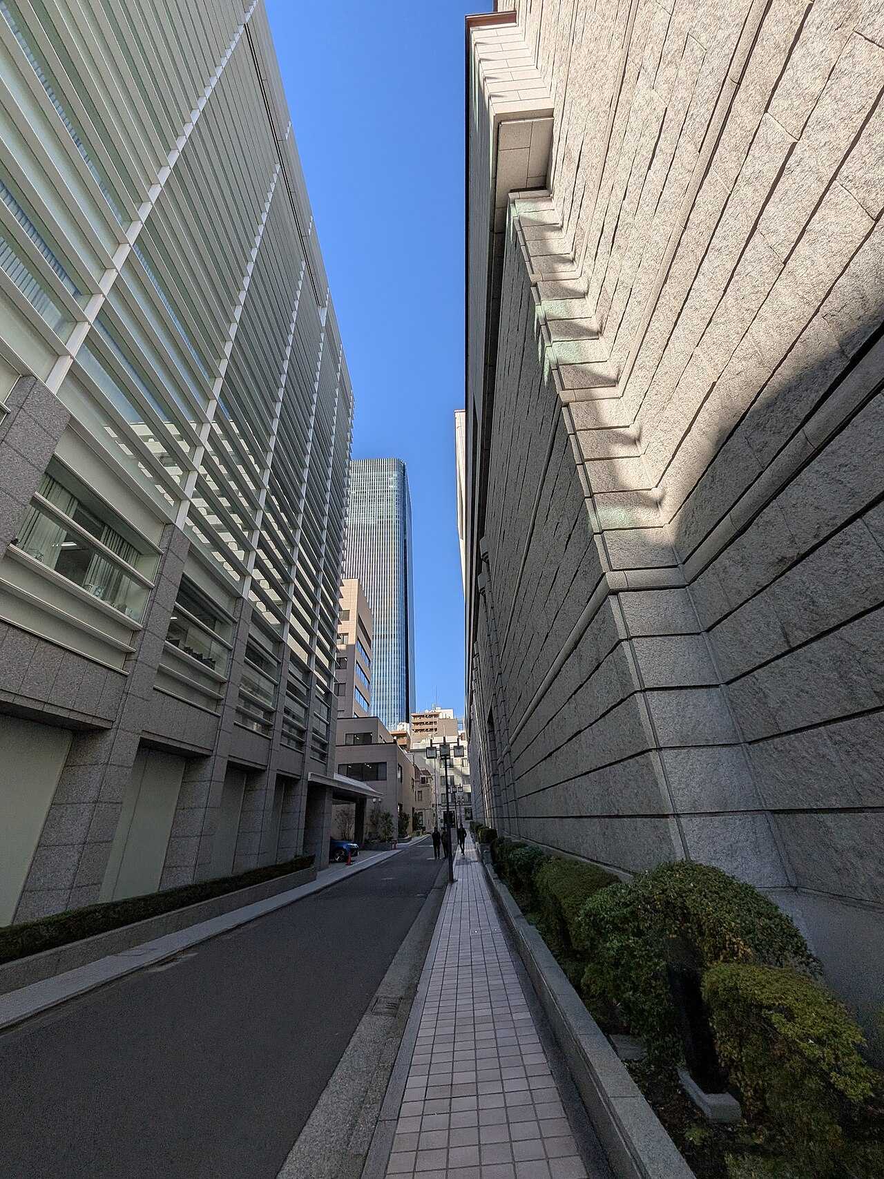A narrow street between a tall stone building and a glass-louvered office building, with a tower behind them under a blue sky
