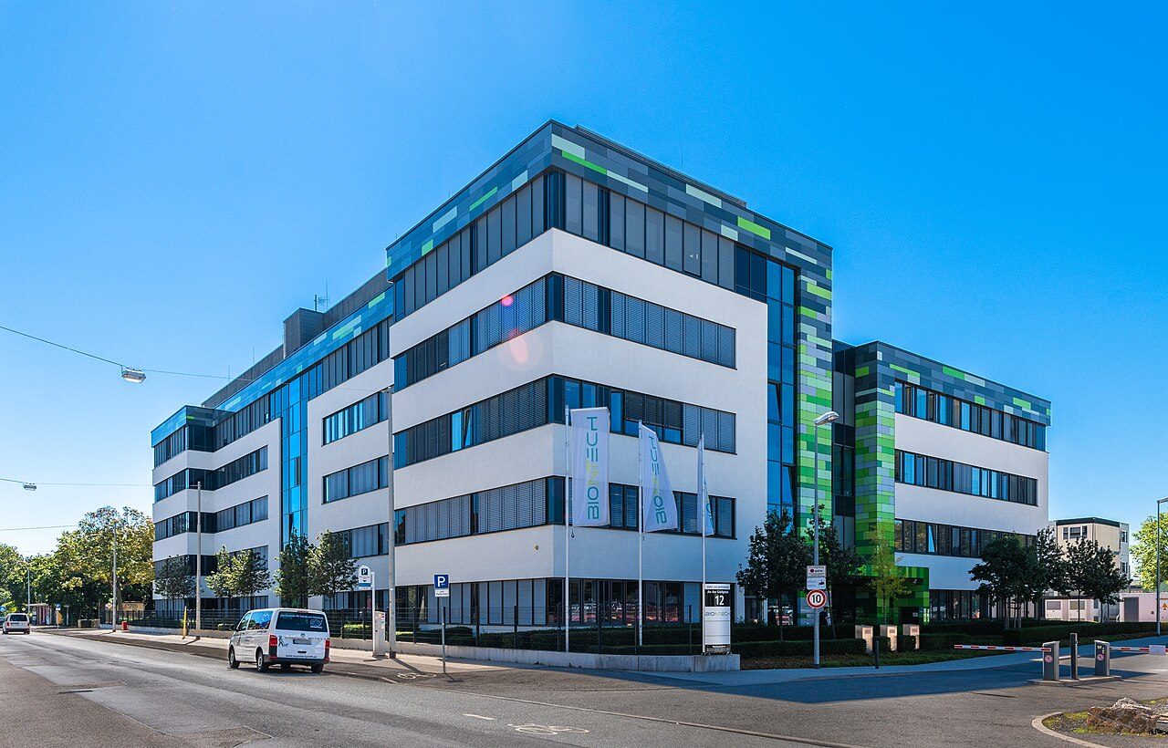 A modern white office building with blue and green glass panels and BioNTech flags out front on a sunny day