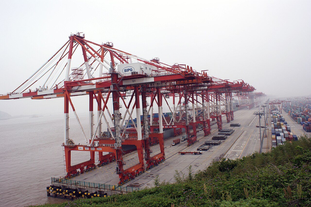 Red container cranes line a long dock at a port, with stacked shipping containers on a misty day