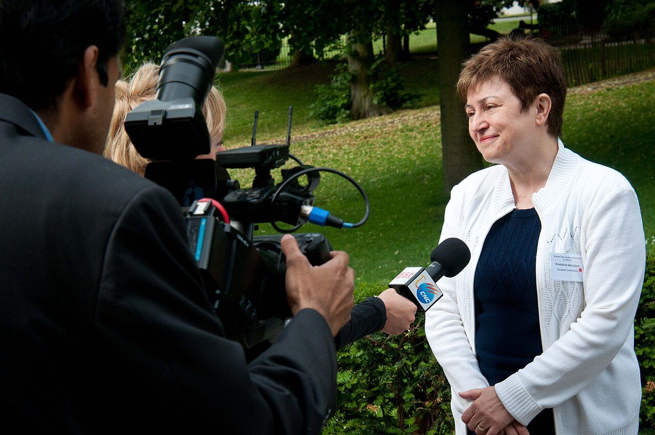 Kristalina Georgieva, in a white jacket, speaks into a microphone held by a TV crew outdoors