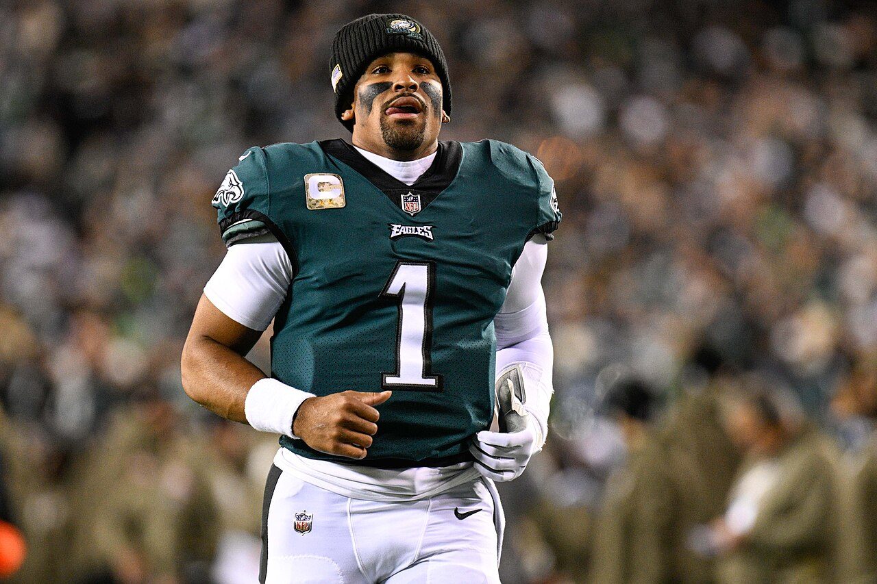 Jalen Hurts, wearing a green Eagles No. 1 jersey and a black knit cap, jogs on the field with a blurred crowd behind him