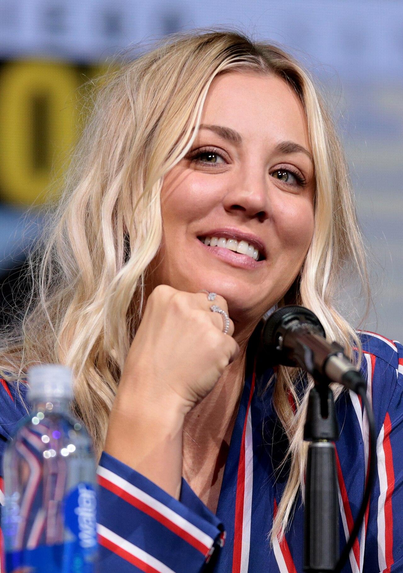 Kaley Cuoco smiles and rests her chin on her fist while seated at a microphone