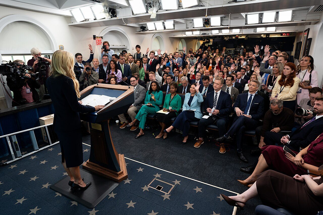 Katie Zacharia, seen from behind at the podium, faces reporters raising their hands in the White House briefing room