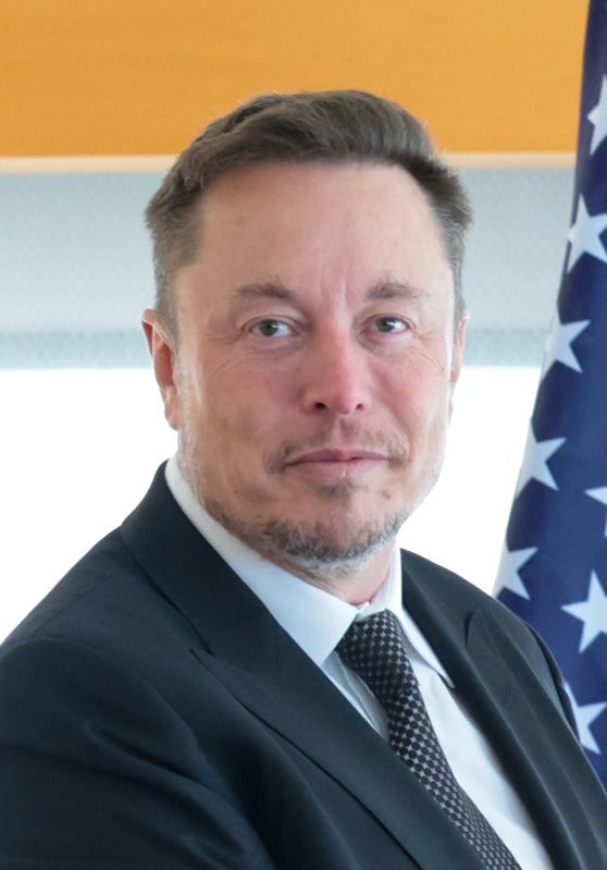 Elon Musk in a dark suit and tie stands in front of an American flag