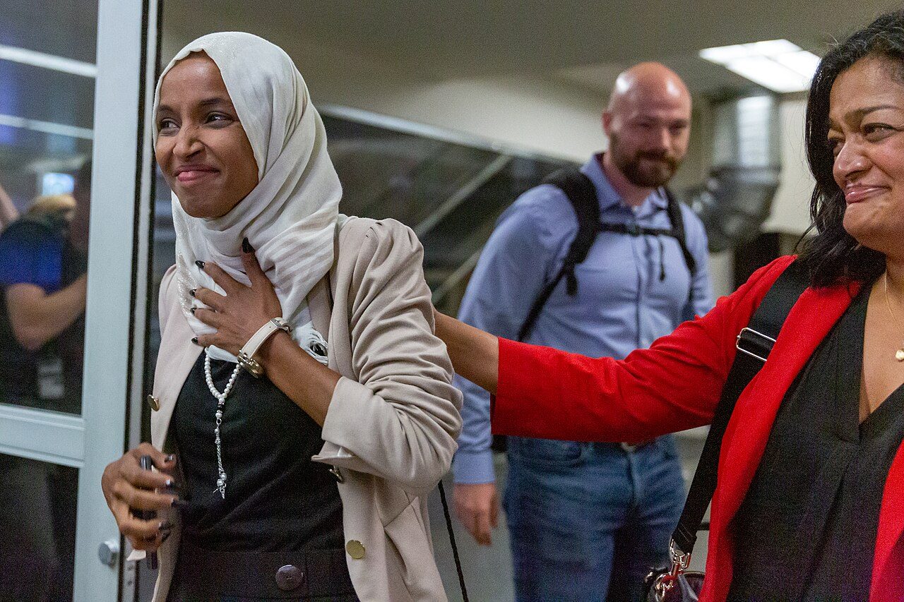 Rep. Ilhan Omar, in a white headscarf and beige jacket, smiles with a hand on her chest as Rep. Pramila Jayapal, in a red jacket, reaches toward her shoulder