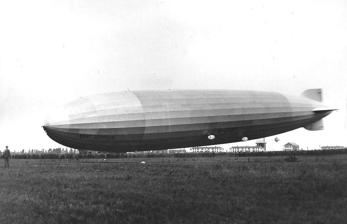 A huge airship with Graf Zeppelin painted on its side floating just above a grassy field, with a man standing at left and low buildings in the distance