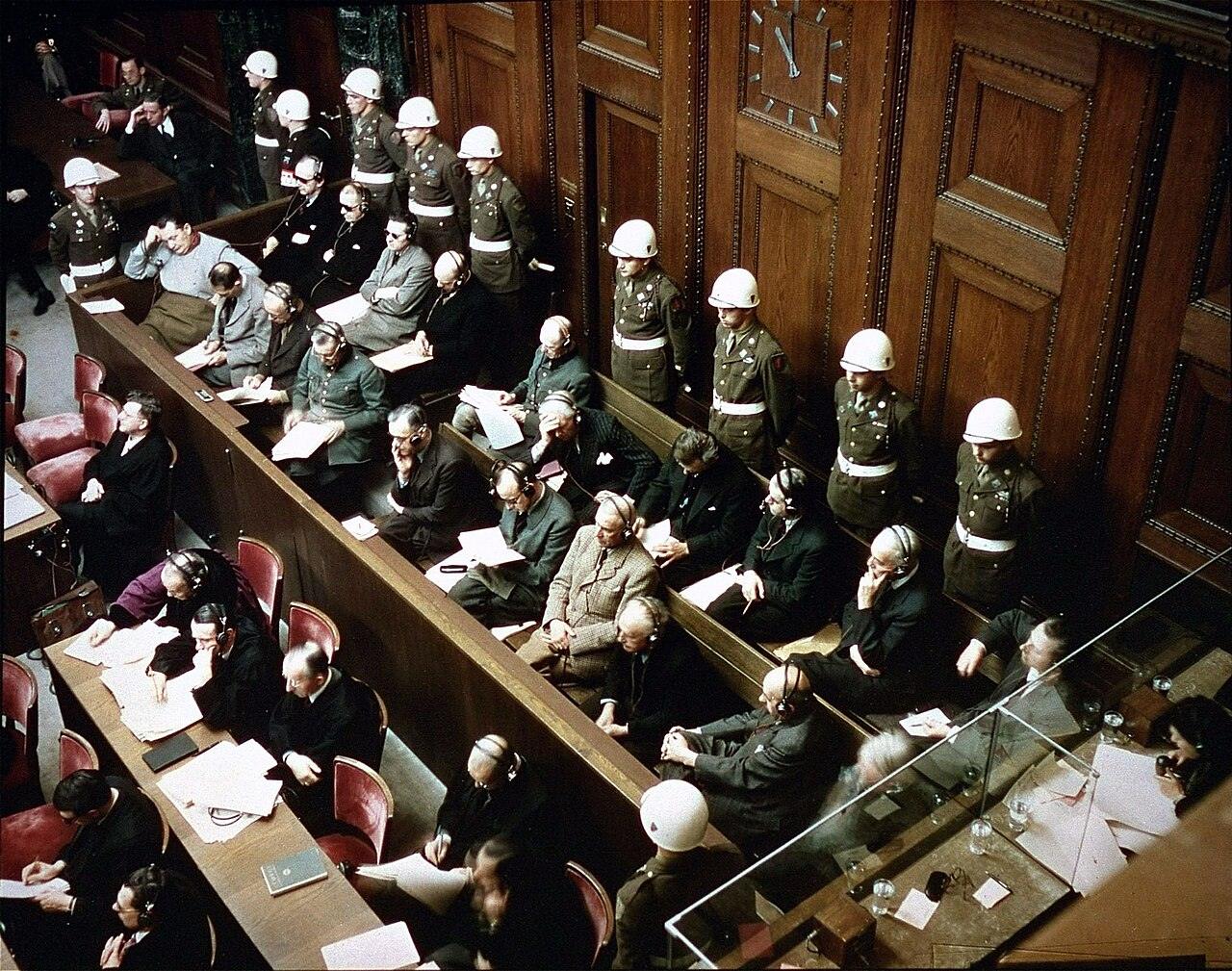 Defendants seated in two rows in the dock of a wood-paneled courtroom, guarded by soldiers in white helmets