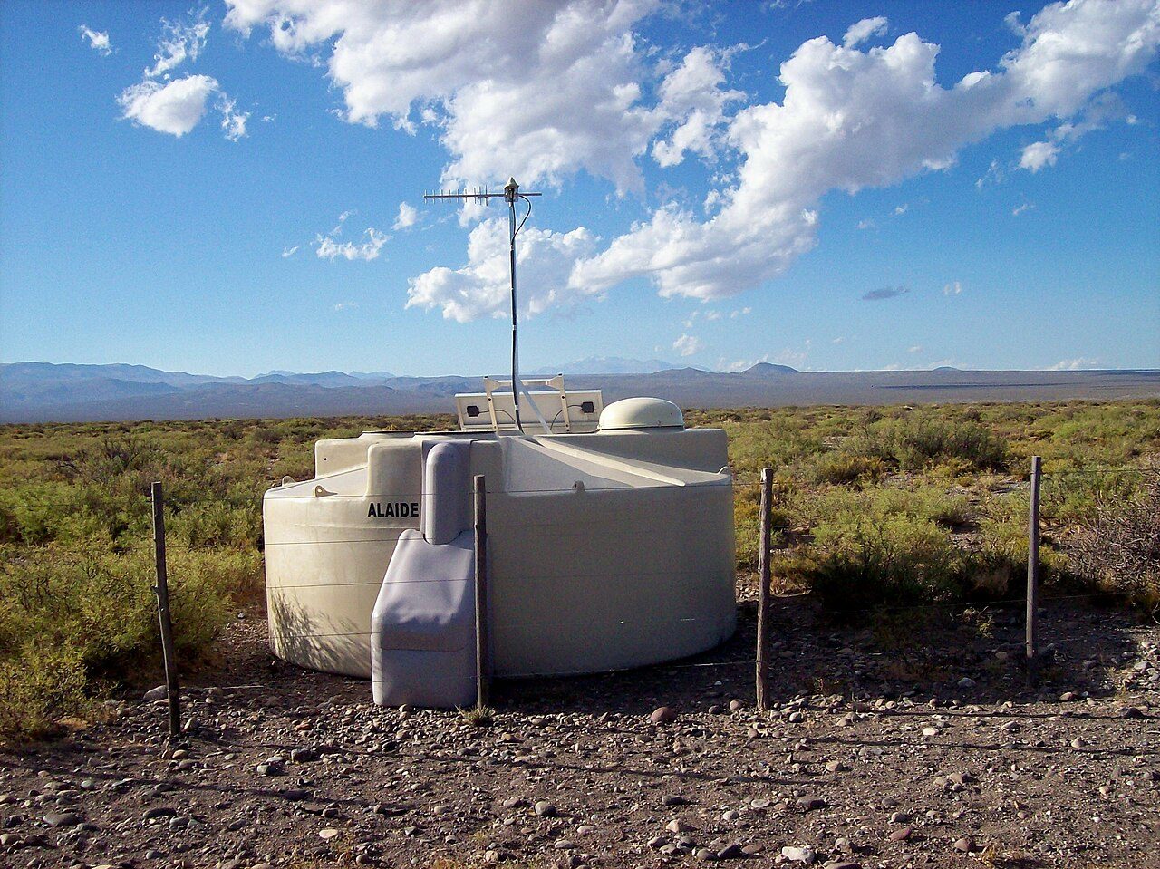 A white cosmic ray detector tank with an antenna in a dry, grassy plain under a blue sky