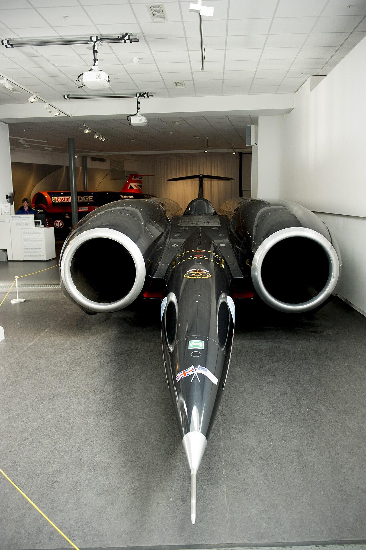 A long black jet-powered car with two large engine intakes and a needle nose on display in a museum hall