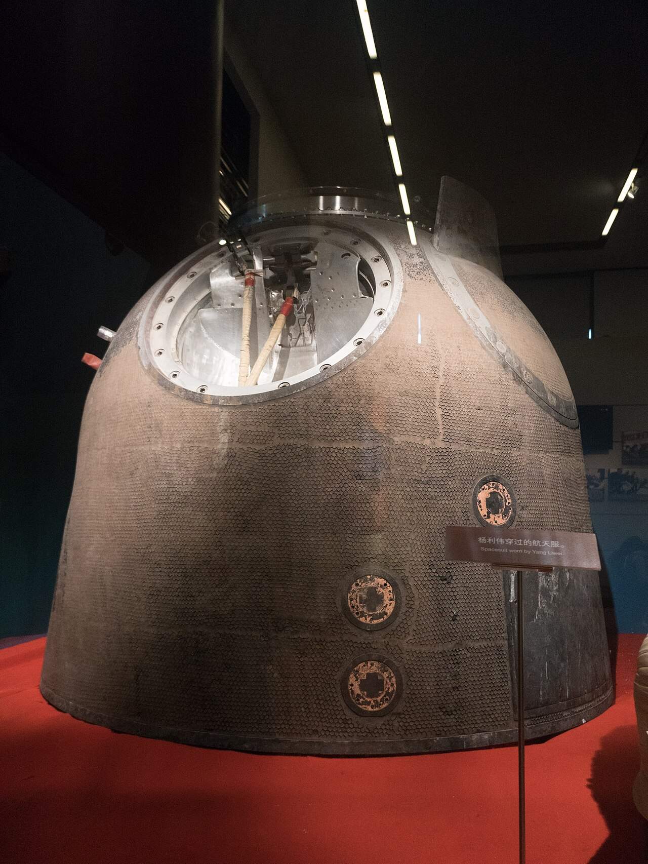 The cone-shaped, scorched brown Shenzhou 5 return capsule on a red stand in a dark museum hall