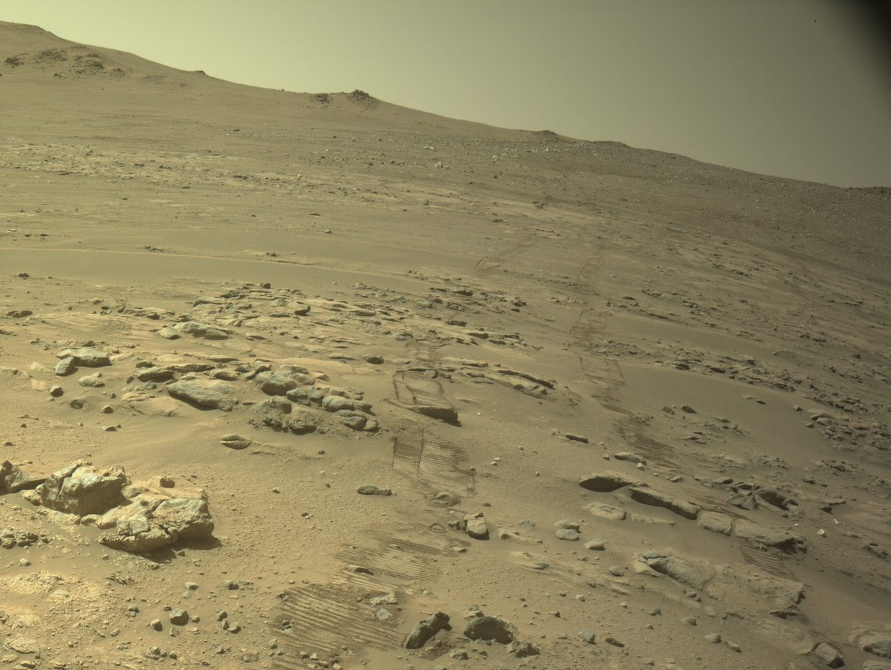 Rover wheel tracks curve across dusty, rocky Martian ground under a hazy sky