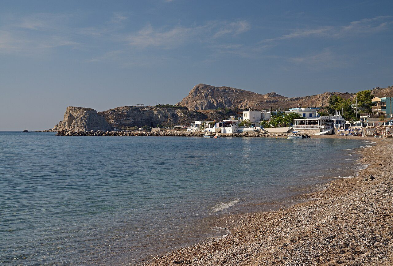 A pebbly beach curves along calm blue water toward a rocky headland and small buildings