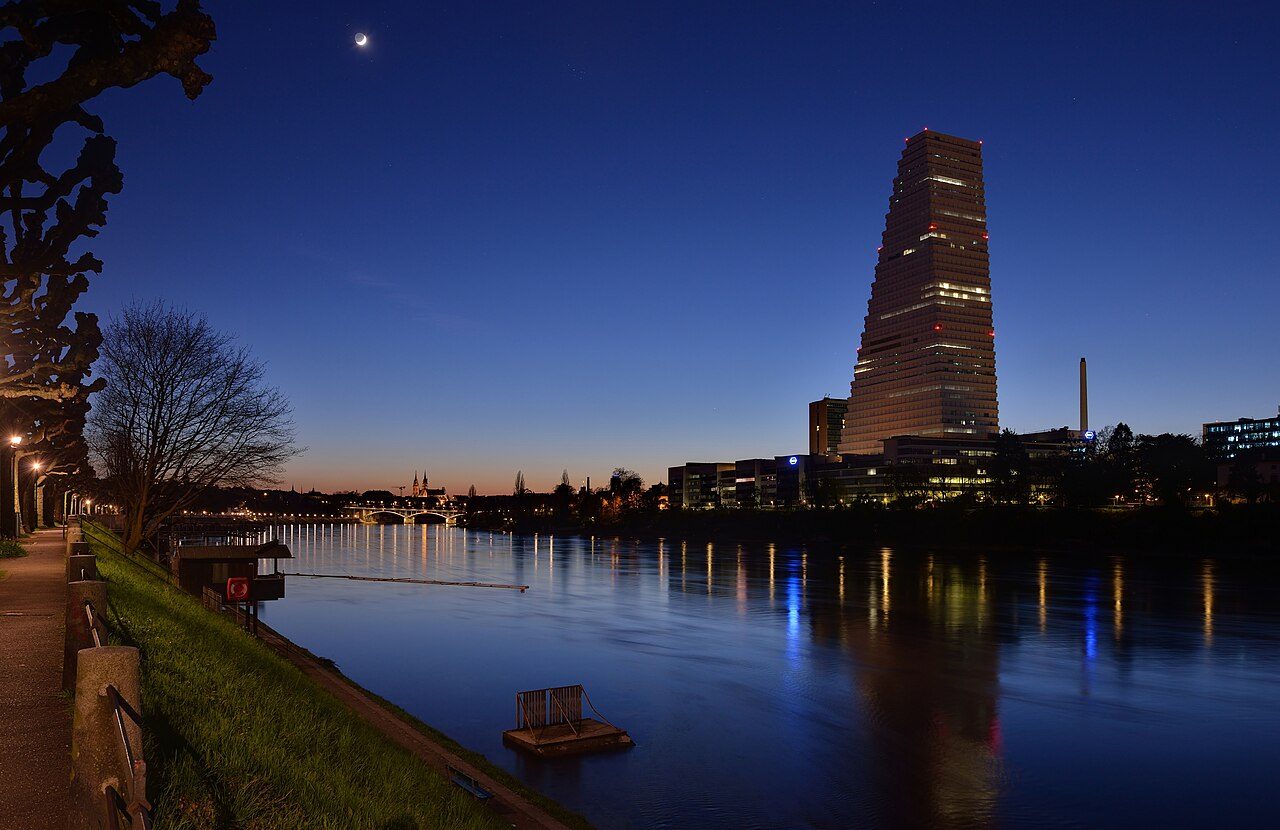 A tall, slender tower lit at dusk beside a calm river with city lights reflected in the water