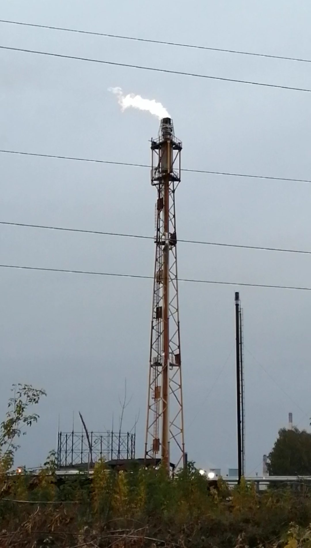 A tall orange lattice flare stack releasing a white plume against a gray sky, with power lines and shrubs in front