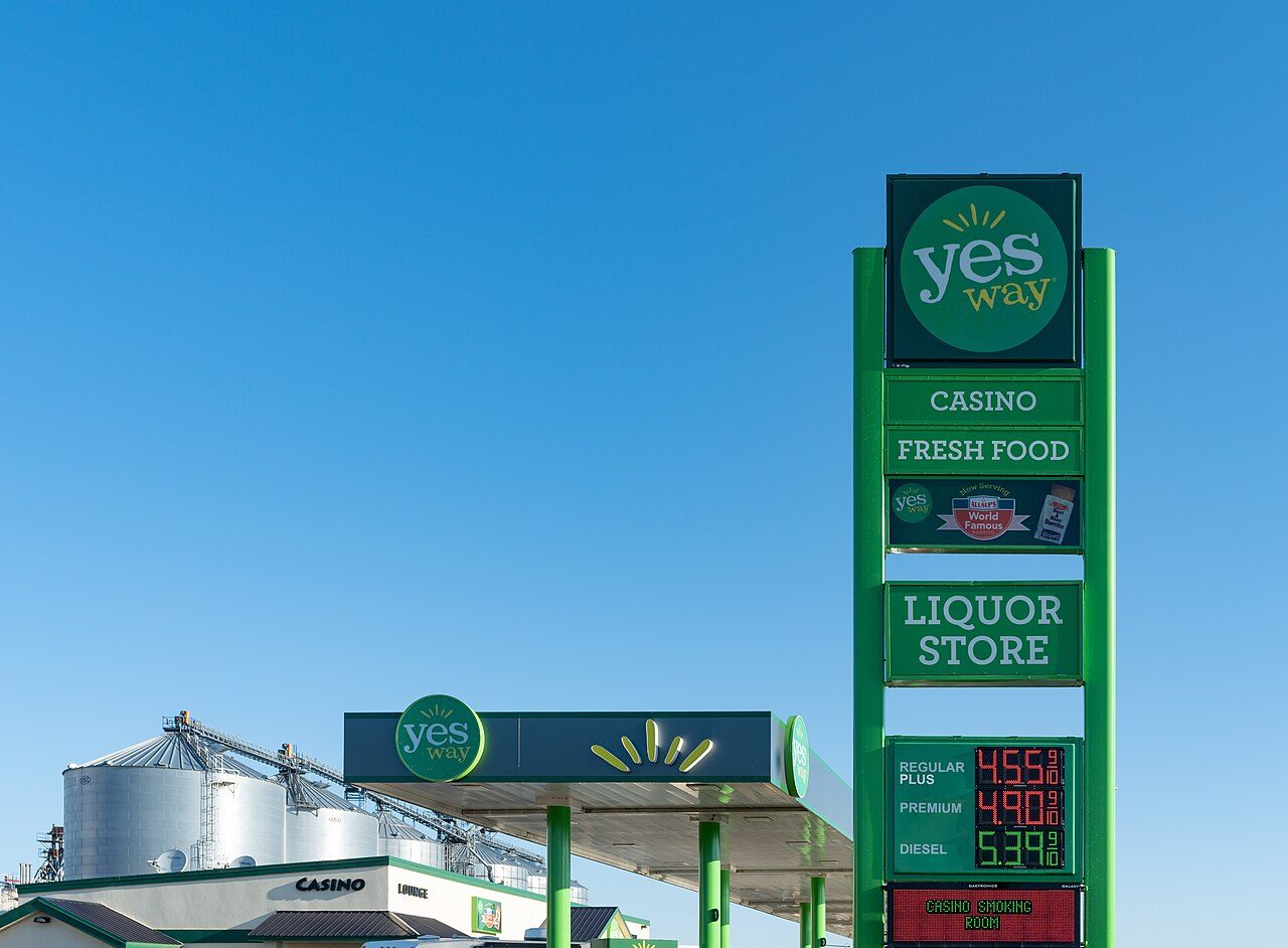 A tall green Yes Way gas station sign showing regular plus, premium and diesel prices next to a fuel canopy under a clear blue sky