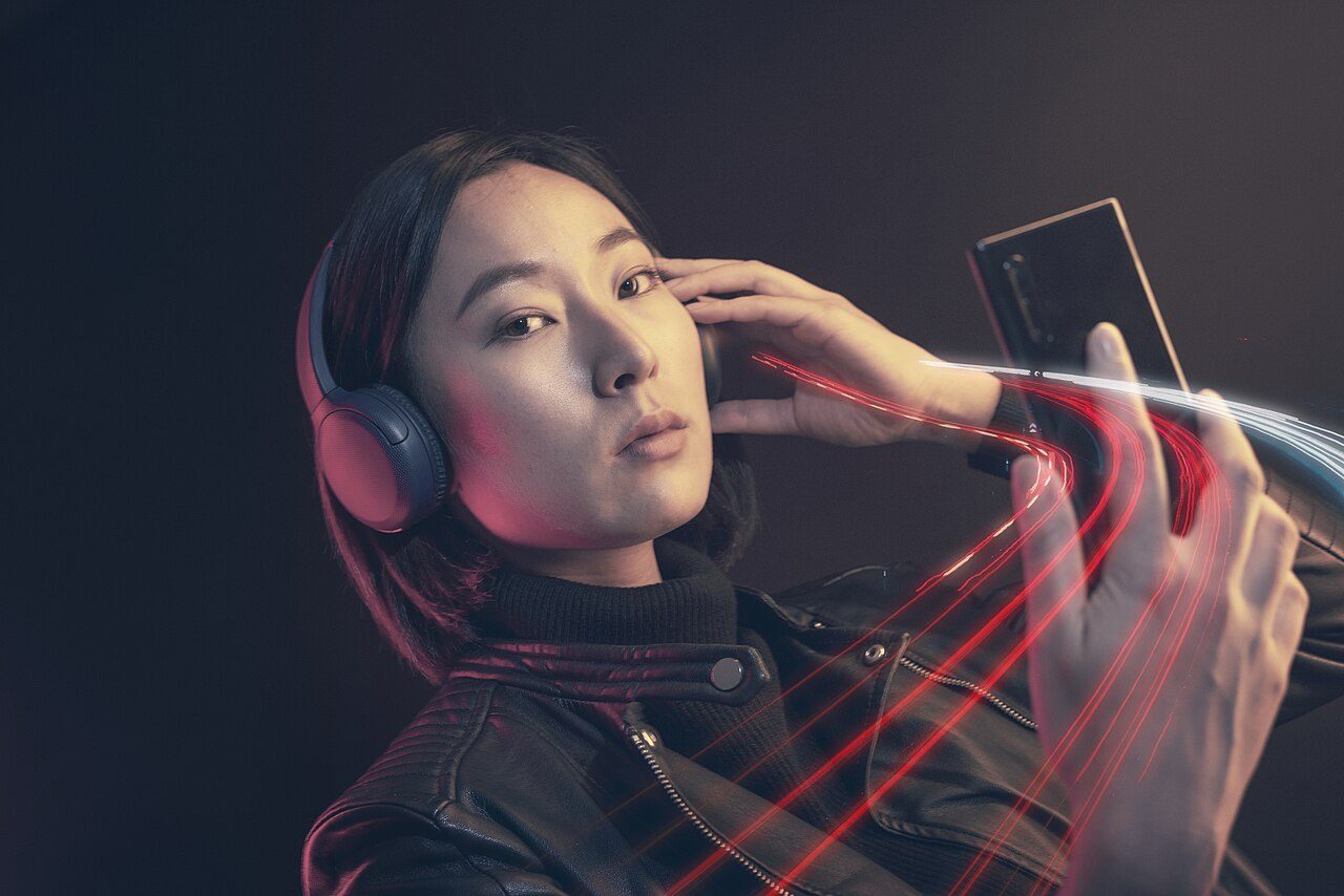 A woman wearing headphones holds a smartphone near her face in a dark room with red light streaks
