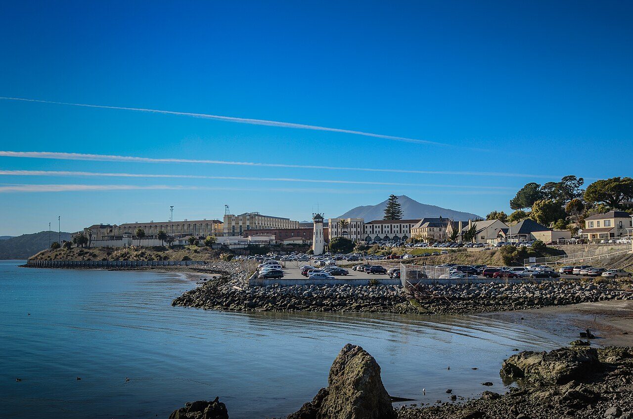Low prison buildings on a rocky shoreline beside calm blue water
