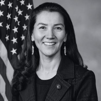 A black-and-white official portrait of Democrat Mary Peltola, smiling, with long dark hair, in front of an American flag