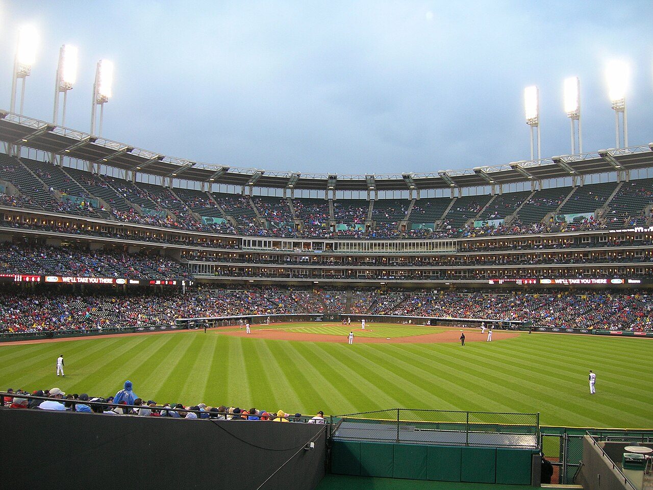 A baseball stadium seen from the outfield stands, with fans in the seats and light towers above a green field