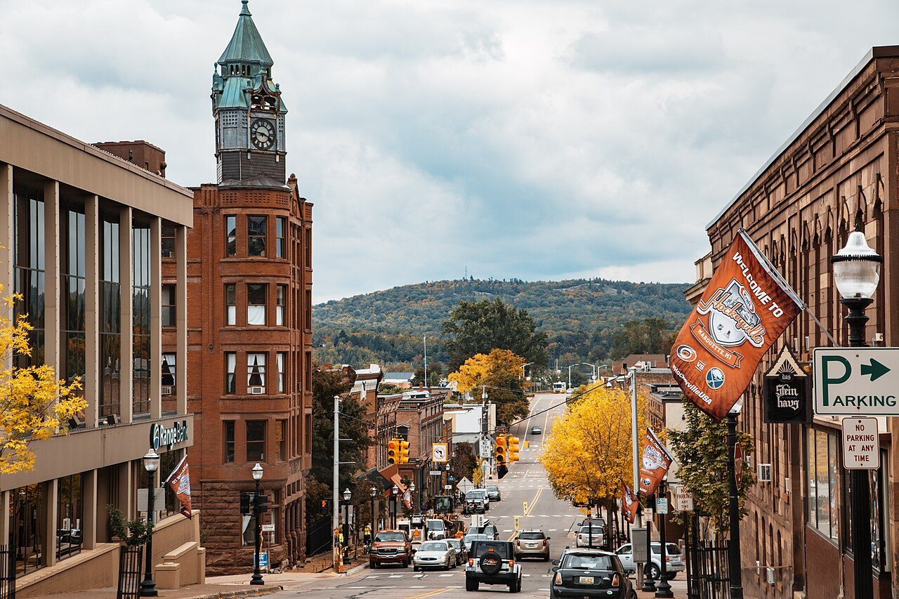 A downtown street of brick buildings with a clock tower runs downhill toward autumn trees and a hill, with a hockey banner hanging from a lamppost