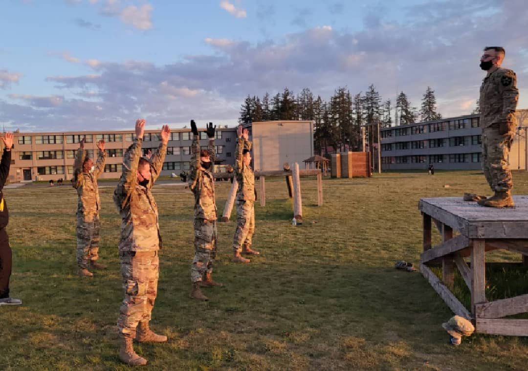 Soldiers in camouflage uniforms stretching on a grassy field as an instructor leads them