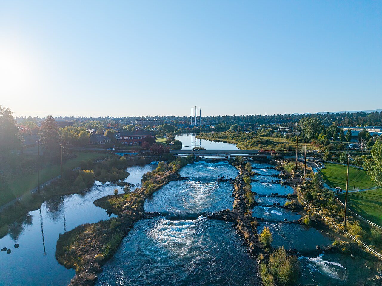 Aerial view of the Deschutes River flowing past green banks and a footbridge in Bend, with three tall smokestacks and the town behind it under a clear sky