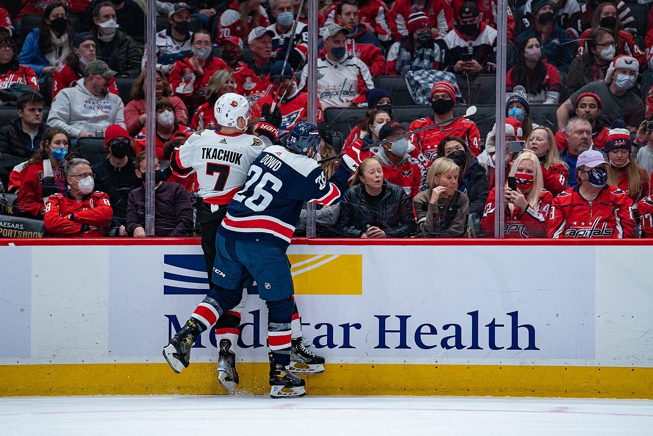 An Ottawa Senators player in a white number 7 Tkachuk jersey is checked into the boards by a Washington Capitals player wearing number 26, with fans behind the glass