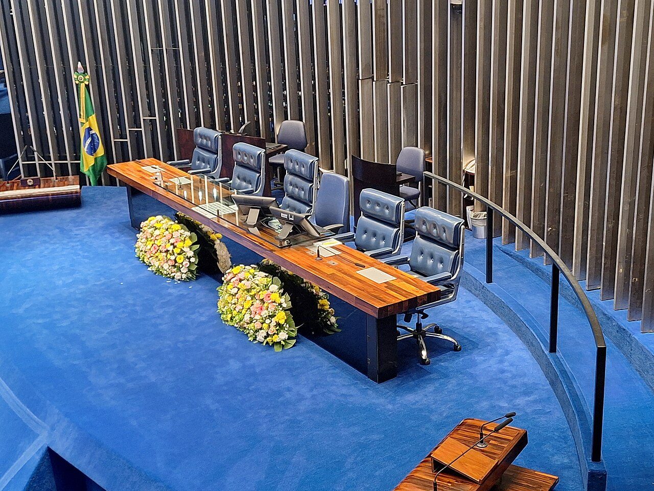 An empty legislative chamber with blue carpet, a long wooden table and rows of black chairs