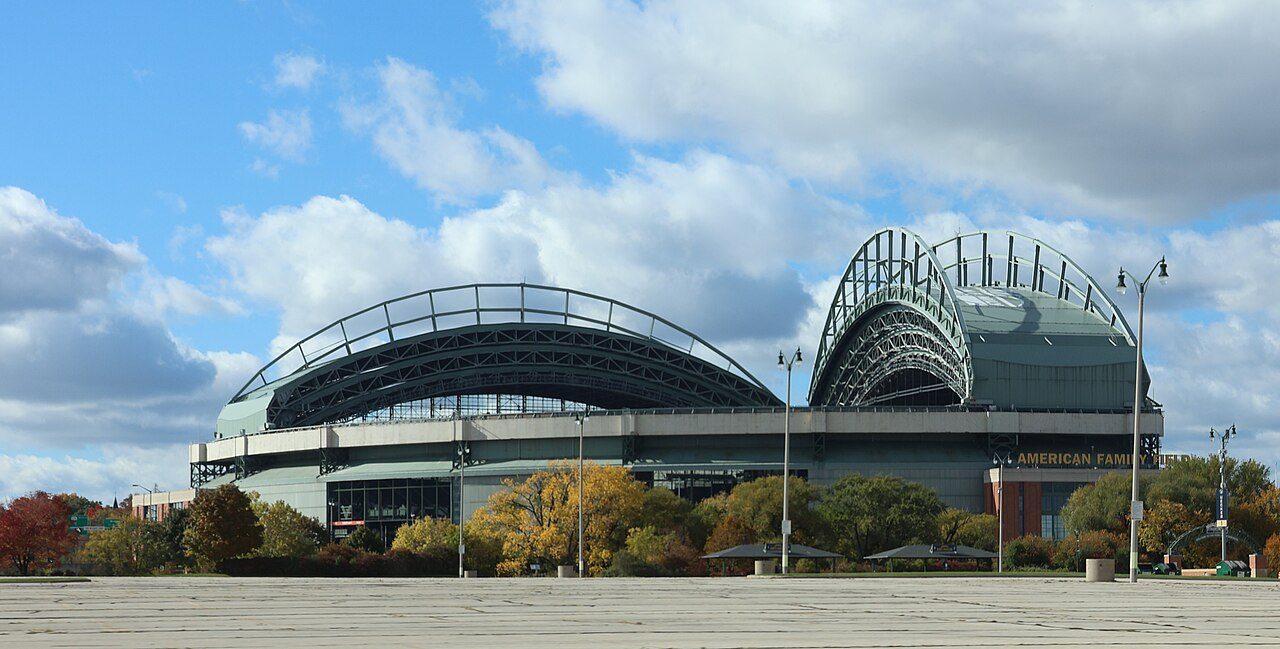 American Family Field in Milwaukee, a ballpark with a large arched steel roof, seen across an empty plaza under a partly cloudy sky with autumn trees in front