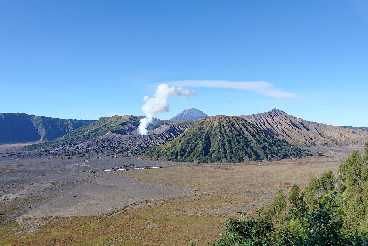 A wide volcanic caldera with a smoking volcano and a tall cone-shaped peak under a blue sky
