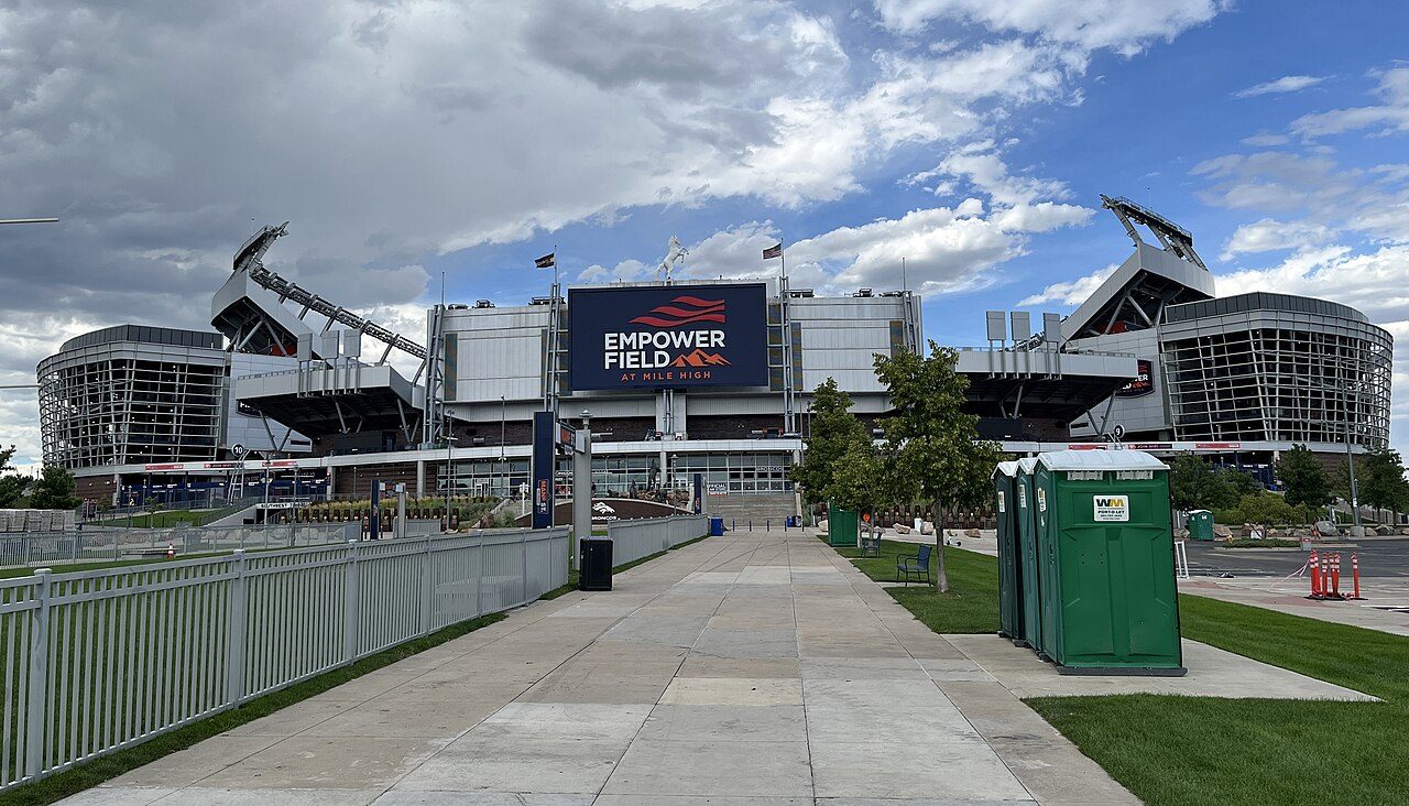 The exterior of Empower Field at Mile High in Denver, with a large video board and a white canopy roof, seen from a wide walkway under a cloudy blue sky
