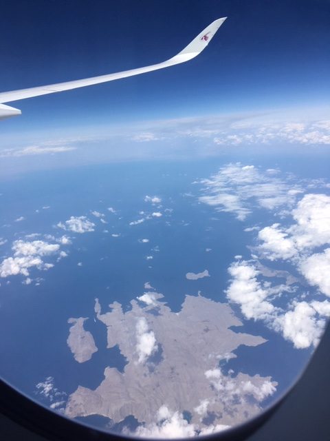 View from an airplane window of a wing above blue sea and scattered white clouds, with a rugged brown coastline below