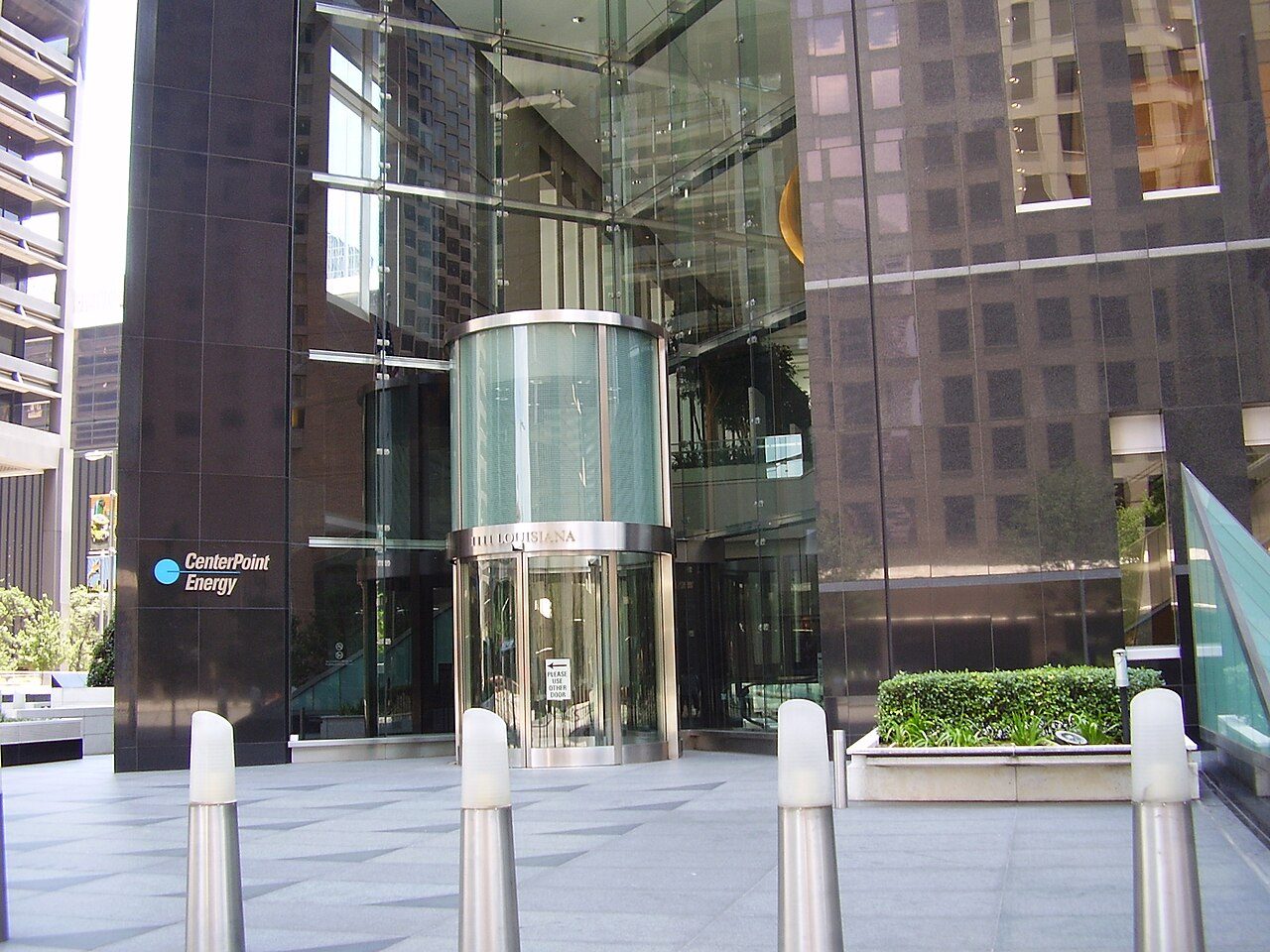 The base of a glass office tower with a round glass entrance and bollards on a plaza