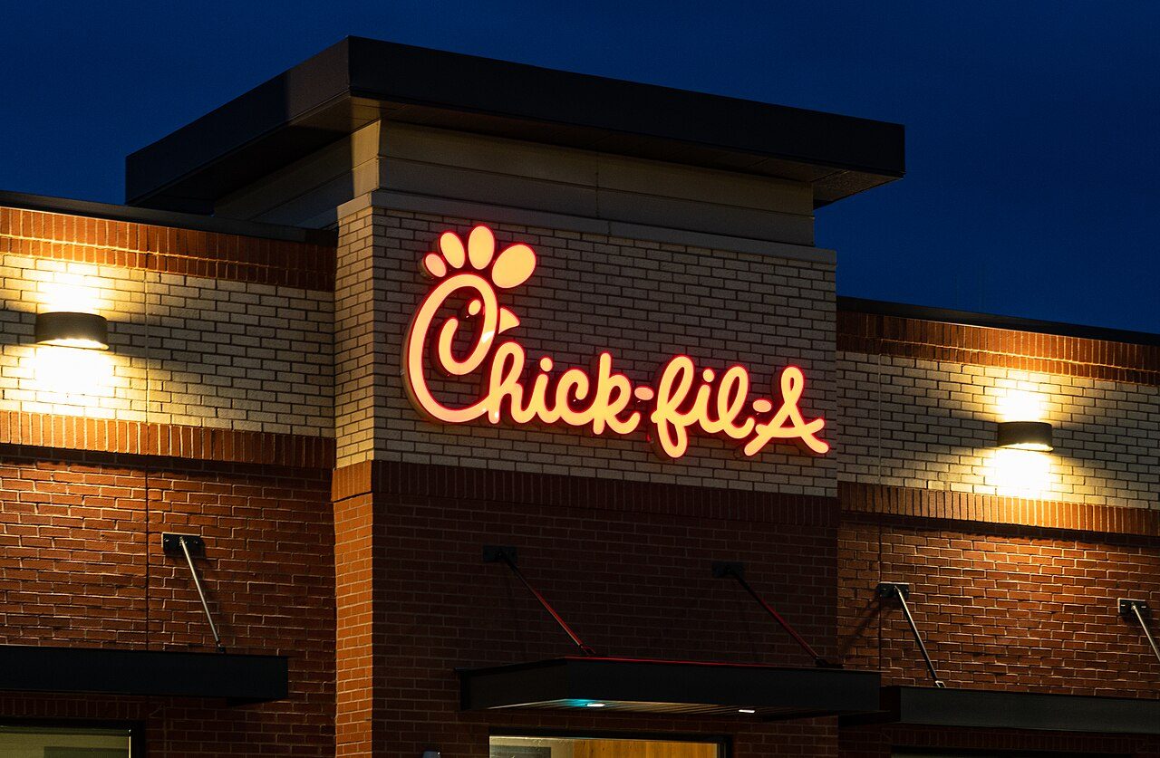 A lit Chick-fil-A sign on a brick restaurant building against a dark blue evening sky