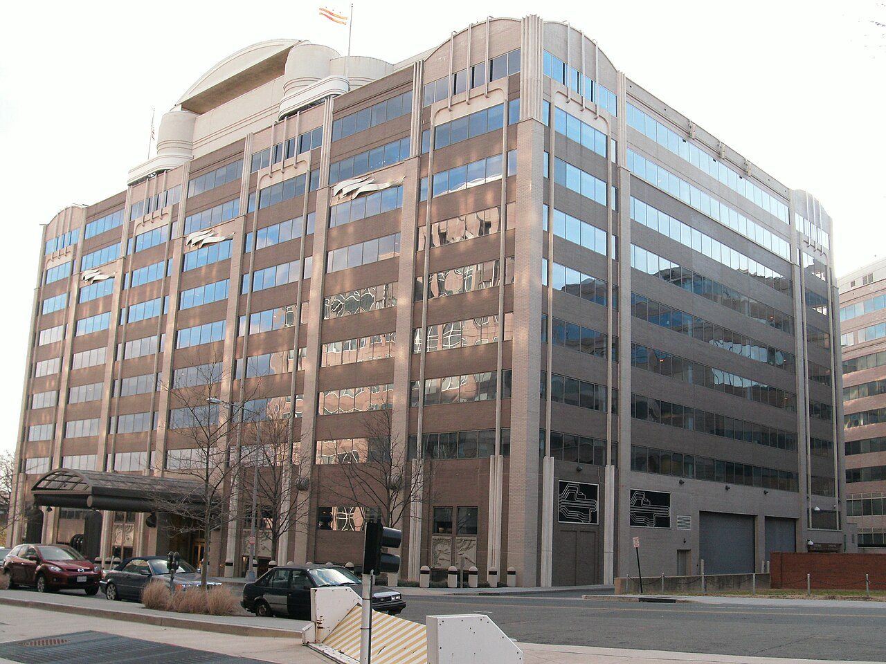 A tall office building with rows of glass windows on a city street, with cars parked in front