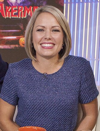 A smiling woman with chin-length blond hair in a blue textured top sits on a TV studio set