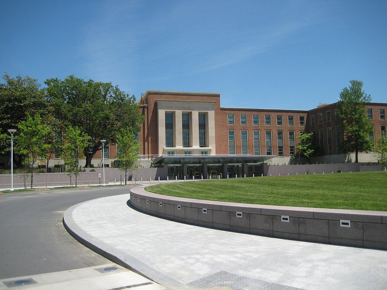 A red-brick and stone government building with tall windows behind a curved concrete walkway and a green lawn, under a blue sky