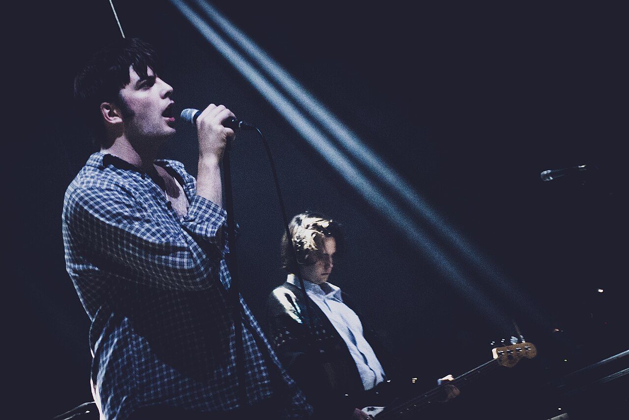 A singer in a checked shirt sings into a microphone under stage lights while a guitarist plays behind him