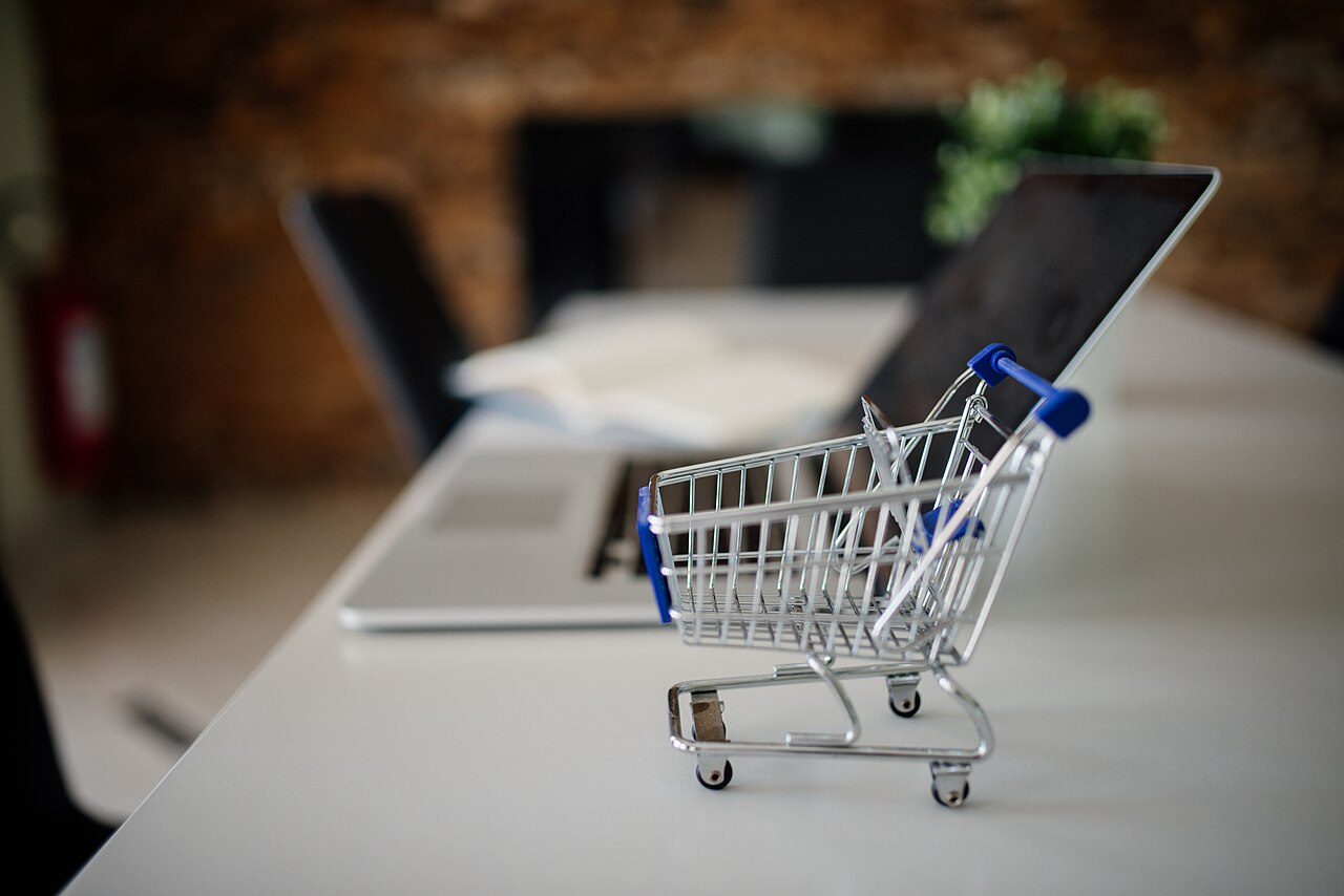 A miniature metal shopping cart sits on a white desk beside an open laptop in an office