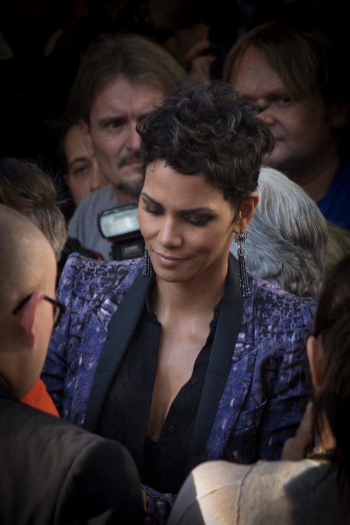 Halle Berry, with short dark curly hair and a sparkly purple jacket, looks down while surrounded by a crowd of people at a film premiere